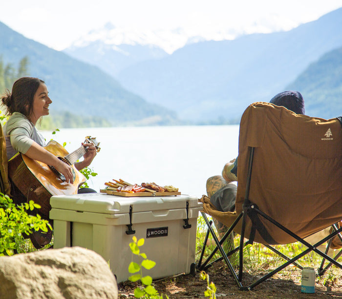 A woman playing guitar next to a man sitting on the Woods Mammoth Folding Padded Camping Chair in Dijon while overlooking water