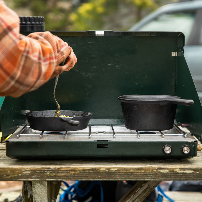 Front view of a person cooking with the Woods Heritage Cast Iron skillet and pot on a portable stove