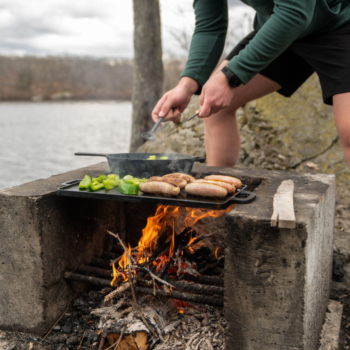 A person cooking vegetables and sausages with the Woods Heritage skillet and griddle over a campfire