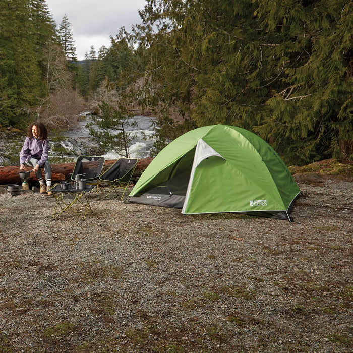 Fully built Woods Cascade Lightweight 2-Person 3-Season Tent next to a woman and two camping chairs on campground