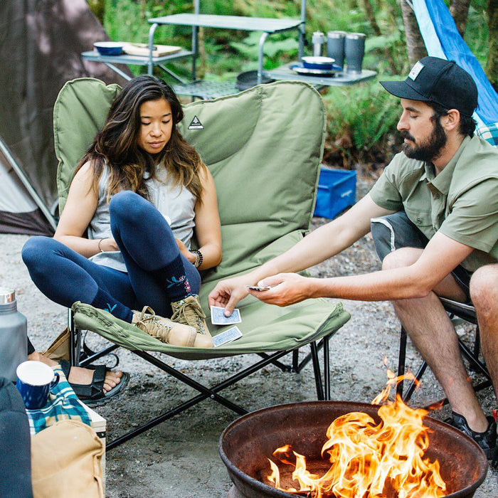 A man handing out cards to a woman sitting on the Woods Mammoth Folding Padded Camping Chair in Sea Spray around a campfire on campground