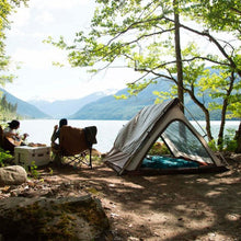 Load image into Gallery viewer, A person sitting on the Woods Mammoth Folding Padded Camping Chair in Dijon next to a tent overlooking the water 