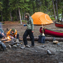 Load image into Gallery viewer, Two people around a campfire with canoes and the Woods Pinnacle Lightweight 2-Person 4-Season Tent in the distance