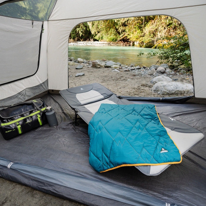 A blanket on top of the Woods Portable Quick Set-Up Adjustable 2-in-1 Camping Cot in Gray next to a duffel bag inside a tent on campground