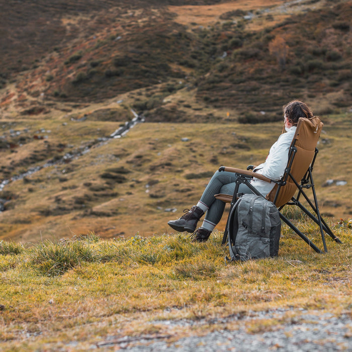 Side view of a person sitting on the Woods Siesta Folding Reclining Padded Camping Chair in Dijon next to a backpack in the outdoors